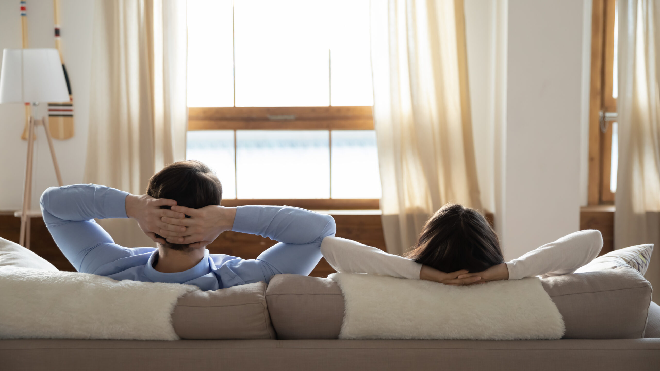 Rear view of young man and woman leaning back relaxed on the couch in front of window with open curtains