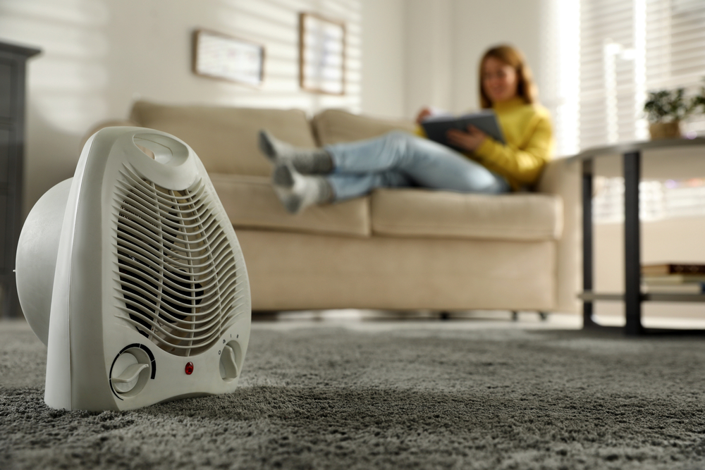 Woman relaxing on the couch, reading a book with an electric space heater in the foreground.