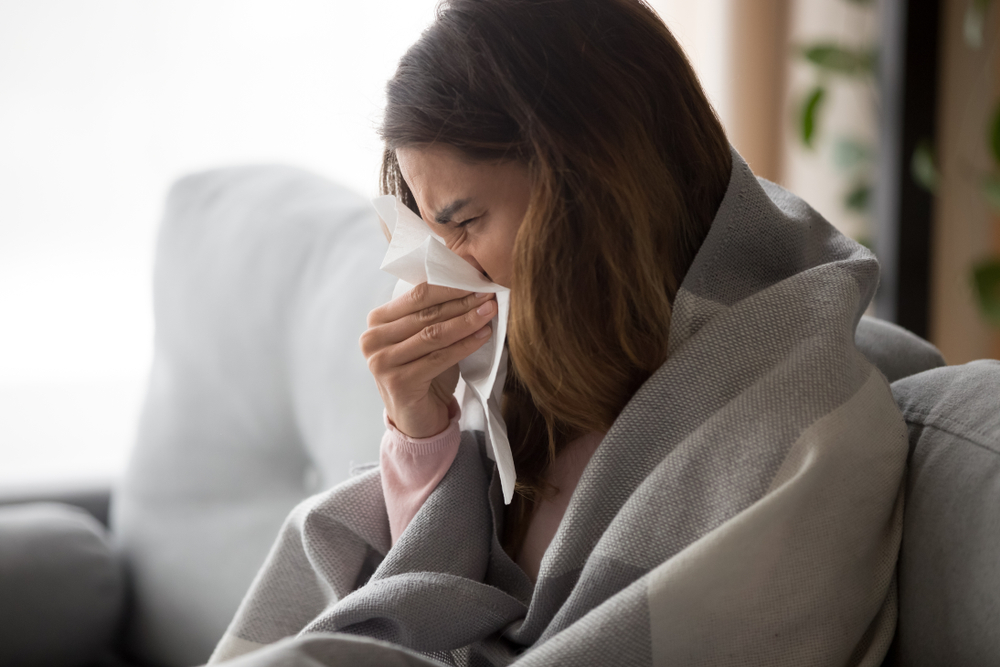 Young woman sitting on a couch, wrapped in a blanket and blowing her nose with a tissue.