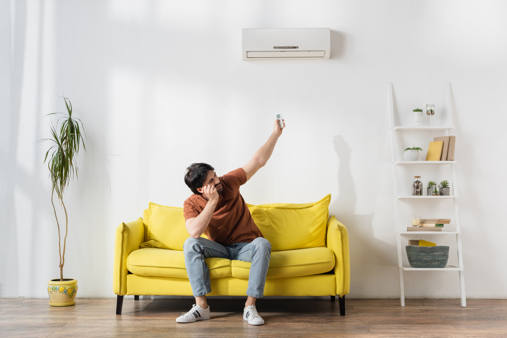 Man sitting on yellow couch, looking annoyed and pointing a remote control at a split system unit.