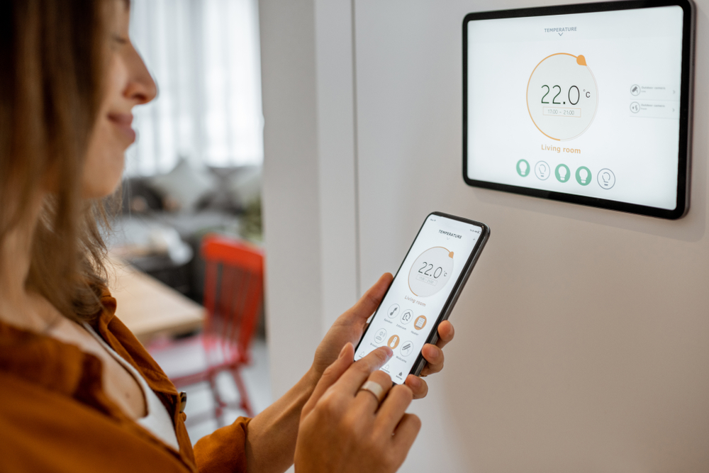 Woman controling temperature of her home using a smart thermostat with phone sychronisation.